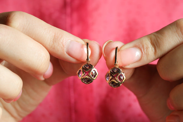 Gold earrings with gemstones held between fingers against a pink background
