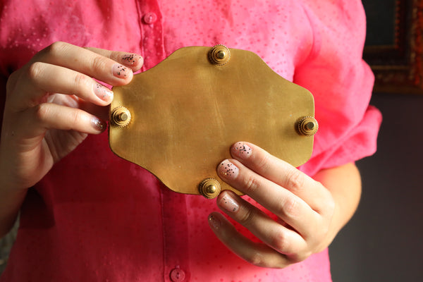 Person holding a gold metal object with four screws against a pink shirt background