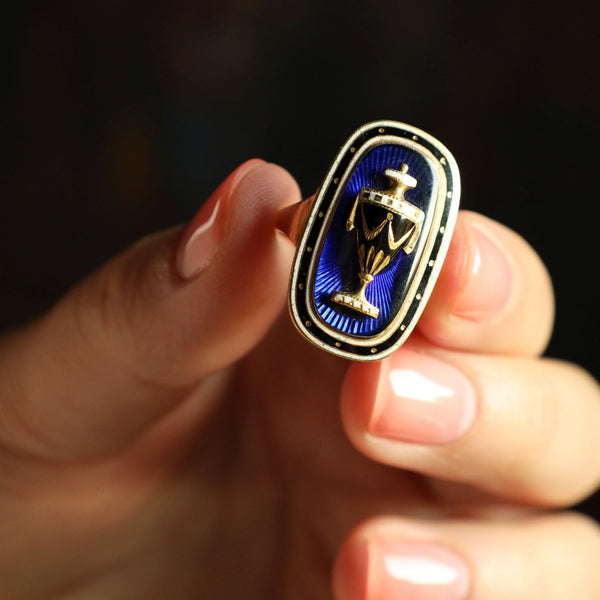 An absolutely fabulous memento moring antique urn ring! This Georgian 18th century ring is a fine example of British sentimental mourning jewelry! Dating back to the year 1793 this panel ring is adorned with a beautiful urn accented in white enamel. Exeptional for it's age, the ring is preserved in outstanding condition with no surface grinding or enamel loss. The rear side of the panel has a clear glass with blond lock of hair and inscription.

Boasting numerous references in antique jewelry books, this wh
