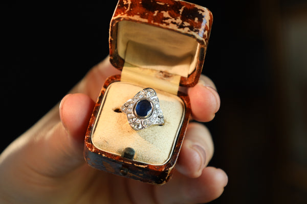 A vintage silver ring with a dark blue oval center stone and clear stones in a decorative halo setting, displayed inside an open, worn reddish-brown wooden ring box held by a hand against a dark background.