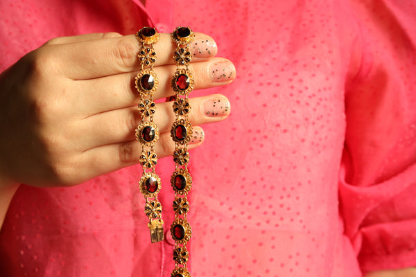 Hand holding a decorative gold and red beaded necklace against a pink background
