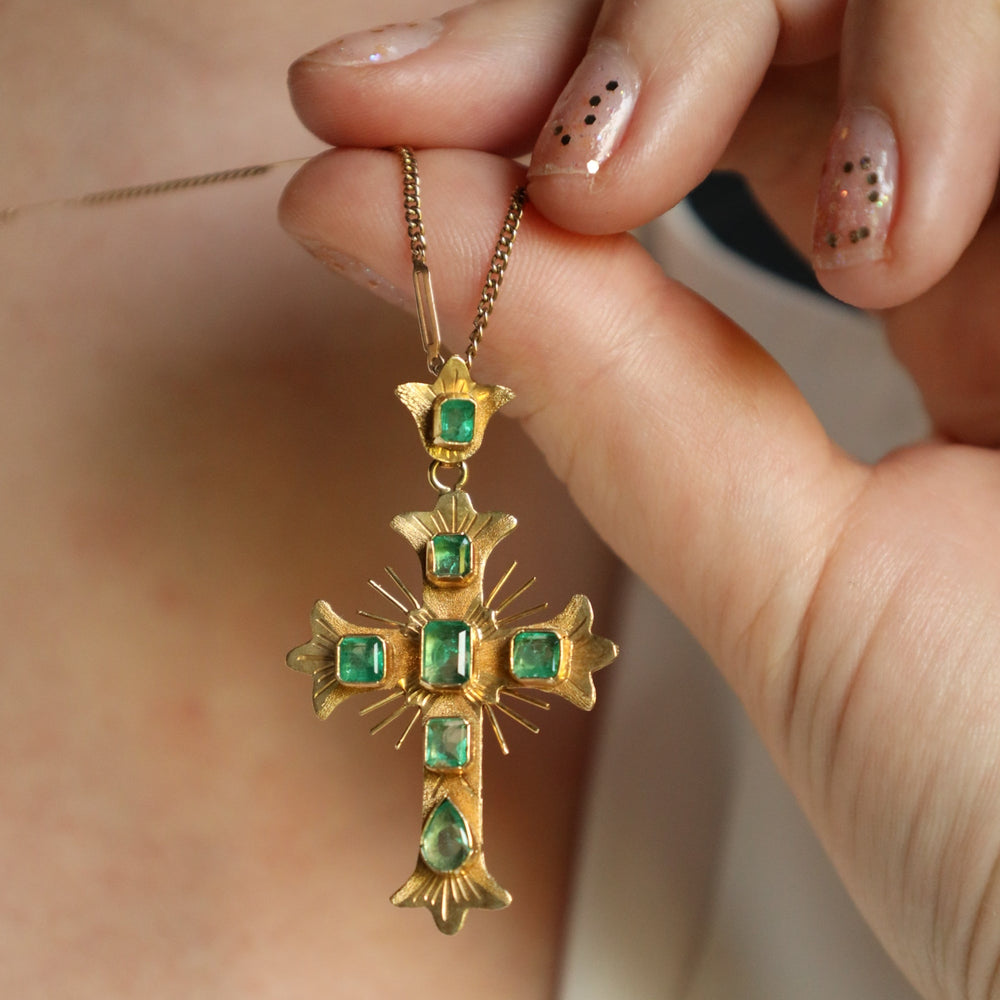 Gold cross pendant with green gemstones held by a hand against a neutral background