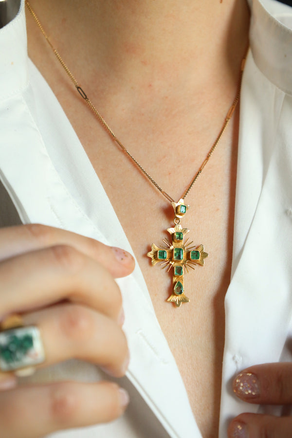 Gold cross necklace with green gemstones worn by a person, close-up on a white background.