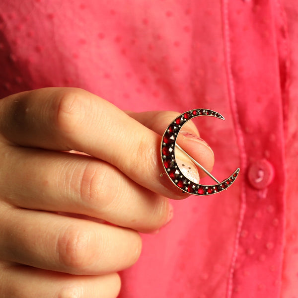 Hand holding a crescent moon brooch with red stones against a pink background