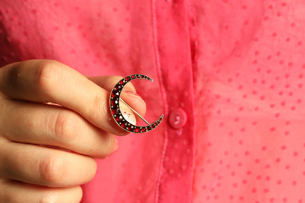 Hand holding a crescent moon brooch with red stones against a pink background
