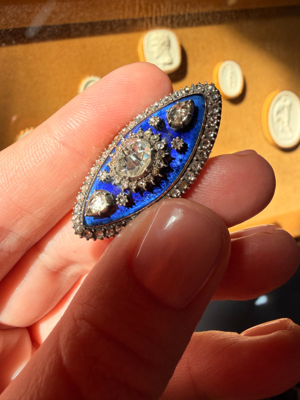 An oval-shaped vintage-style brooch or pin with a vivid blue enamel center, surrounded by silver-toned metal and small clear stones, held between fingers against a warm brown display background.