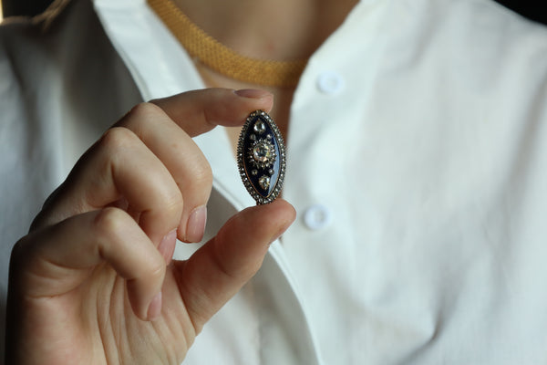 A close-up of a hand holding a small, oval-shaped silver ring with a dark blue enamel background and a central gold-toned floral motif.
