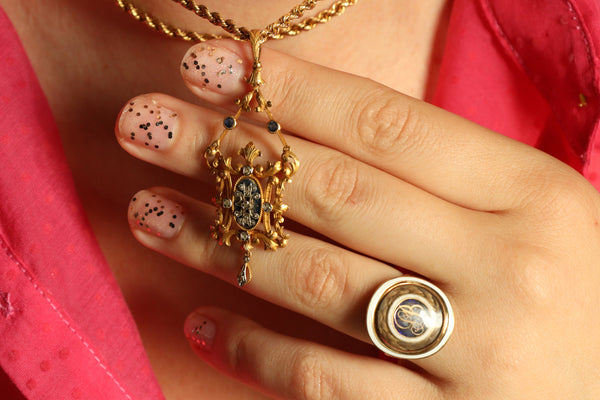 Close-up of a hand wearing a gold ring with a pendant, against a pink background.