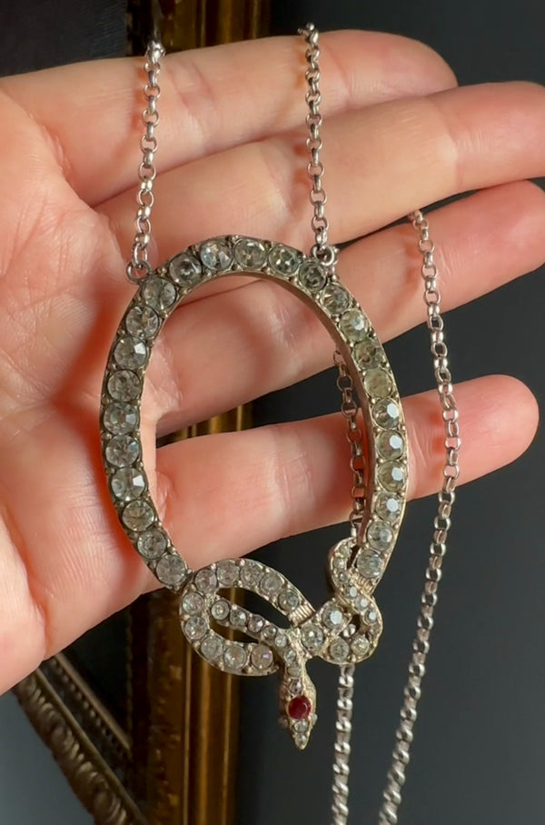 Hand holding a decorative snake-shaped pendant with gemstones on a chain.