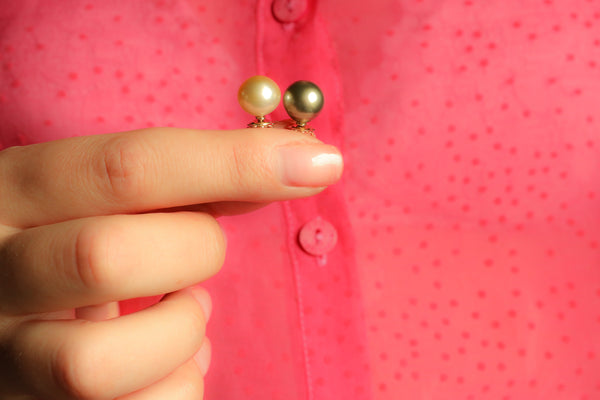 Gold pearl earrings held by a hand against a pink background