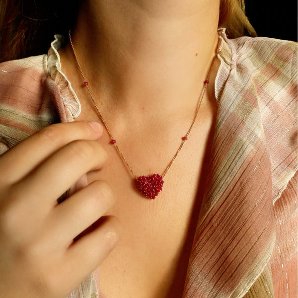 Person wearing a necklace with a red heart pendant, close-up of neck and hand.