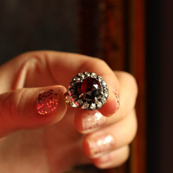 Close-up of a hand holding a diamond ring with a red garnet stone against a blurred background
