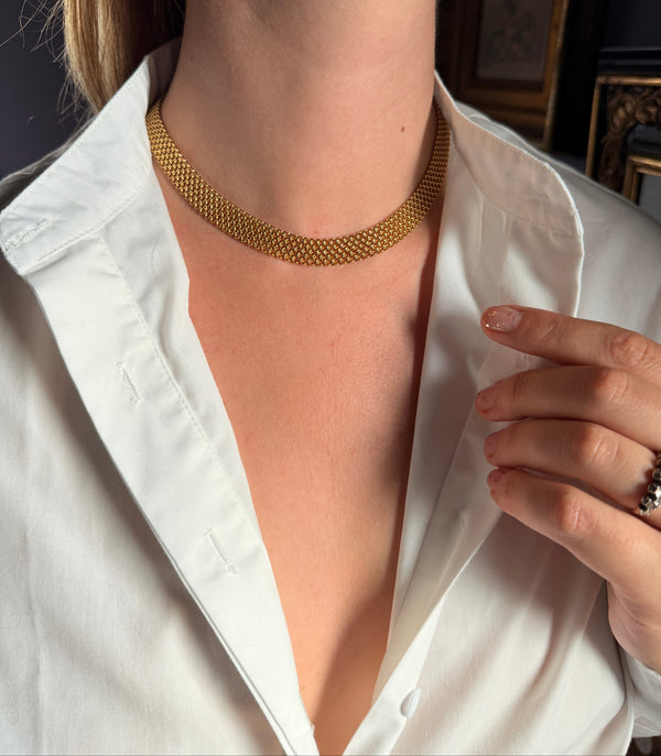 A close-up of a woman wearing a gold mesh choker necklace with a white collared shirt, showing the necklace on her neck.
