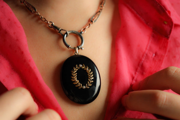 Close-up of a necklace with a black pendant featuring a gold laurel wreath design, worn by a person in a red shirt.