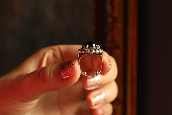 Hand holding a decorative ring with a dark stone and gemstones against a blurred background