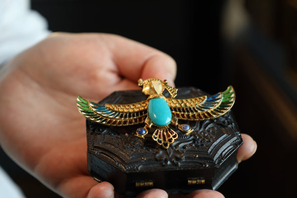Decorative object with a gold bird design and turquoise stone held by a hand against a blurred background