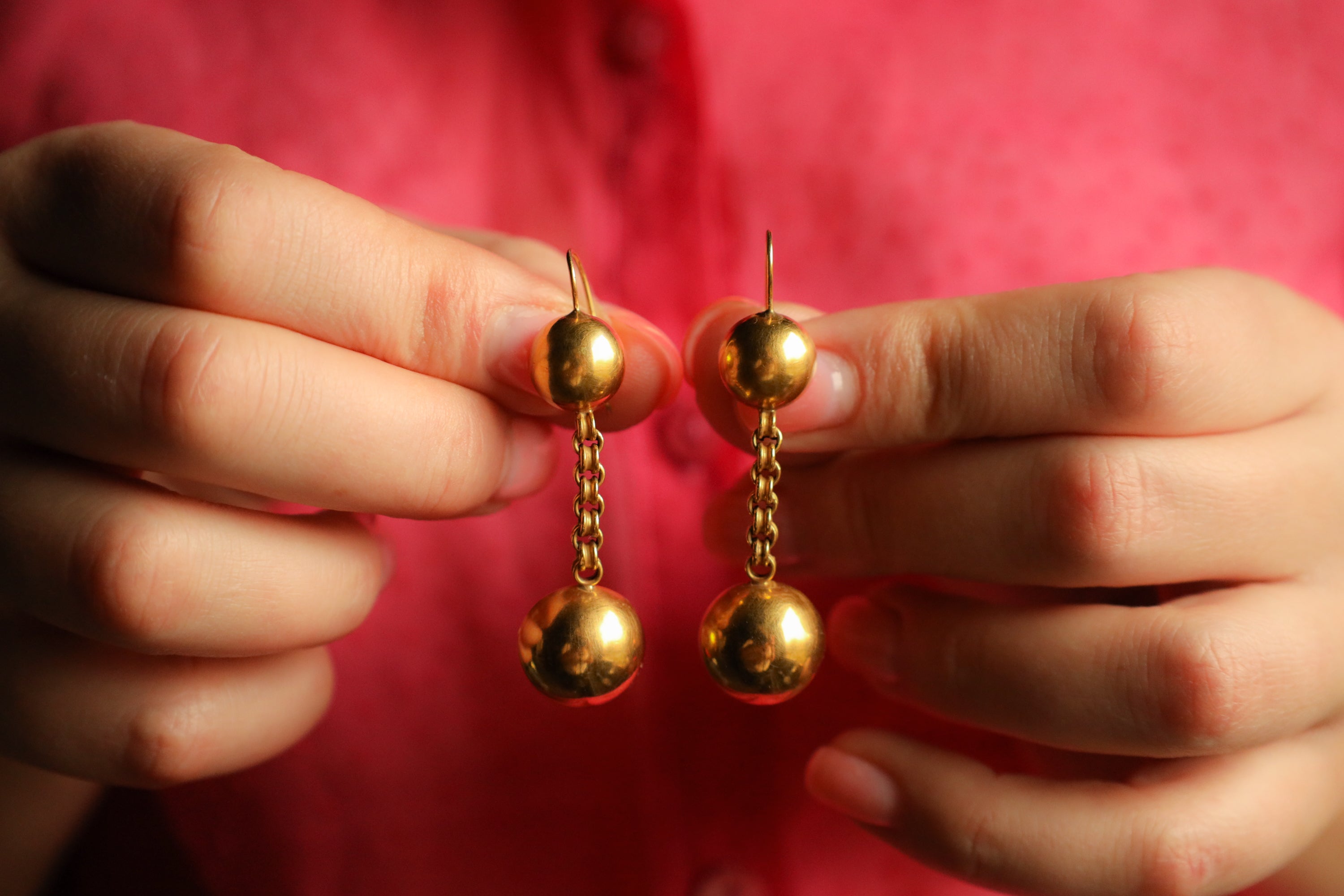 Gold Victorian earrings held between two hands against a red background