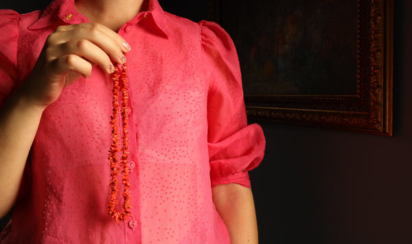 Person wearing a red blouse holding an orange beaded necklace against a dark background