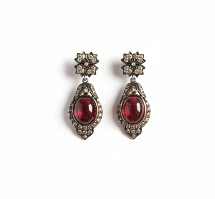 A pair of vintage-style drop earrings featuring oval red gemstones in ornate silver-toned settings with small clear stones, shown on a plain white background.
